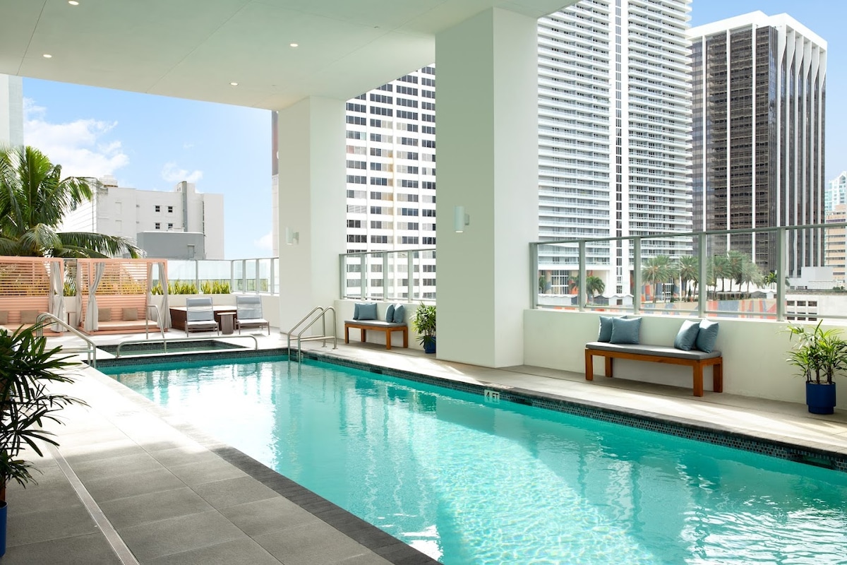 An inviting outdoor pool area is framed by modern architecture and features clear blue water. Lounge seating and small tables are arranged alongside the pool, complemented by potted plants, creating a relaxing environment in a downtown setting.