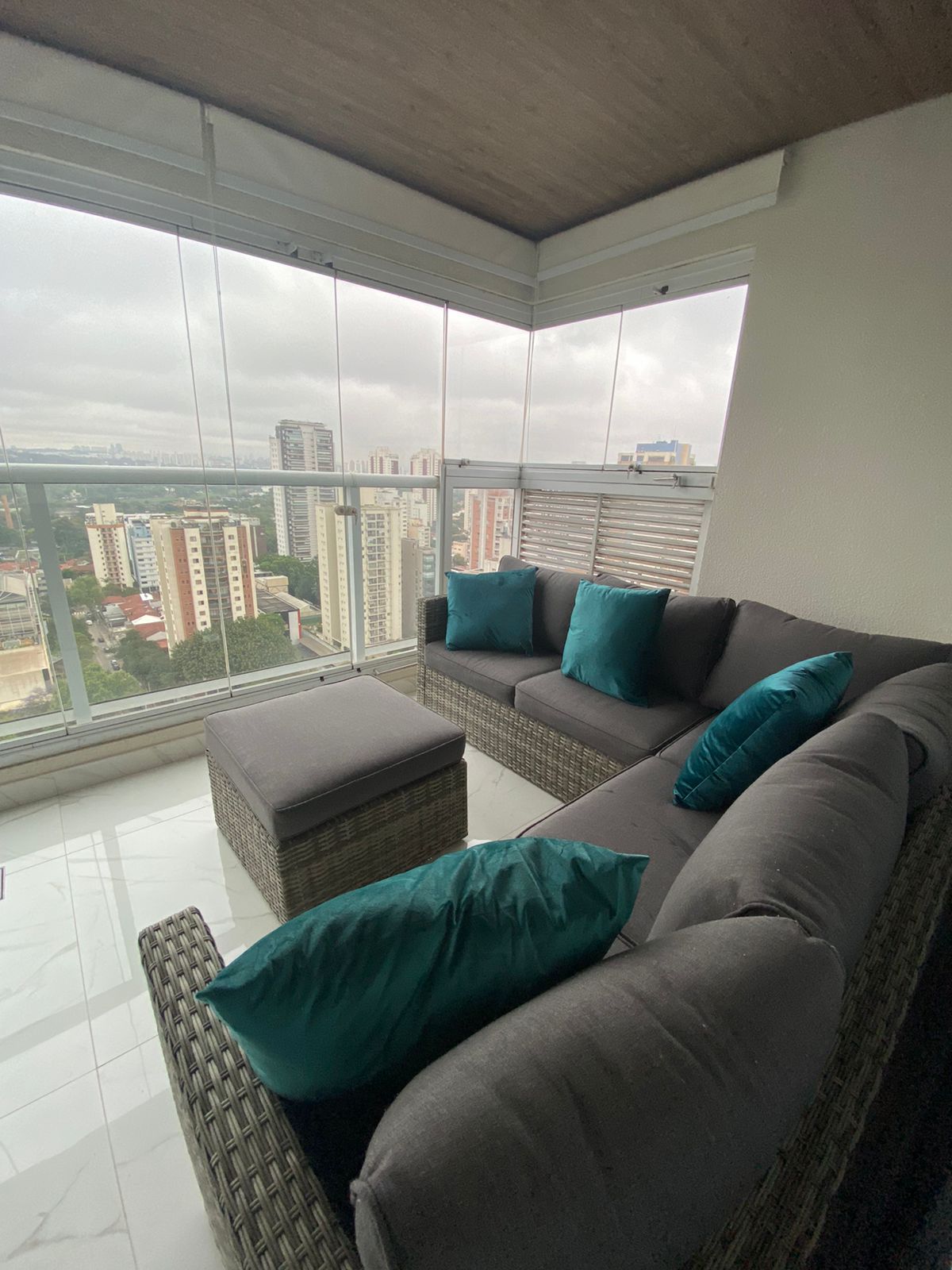 A cozy sitting area is presented on a glass-enclosed balcony, featuring a gray sofa and a matching ottoman. Accent pillows in teal add color, while an expansive view of the cityscape is visible through large windows.