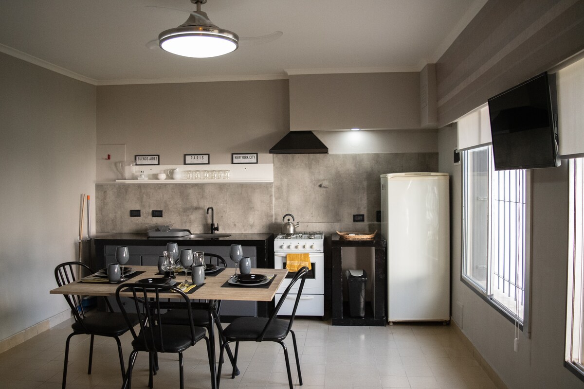 The kitchen and dining area feature a modern design, with a dining table set for six. A sleek black countertop is complemented by a white refrigerator and a stove. Natural light fills the space through window blinds, enhancing the clean, functional layout.
