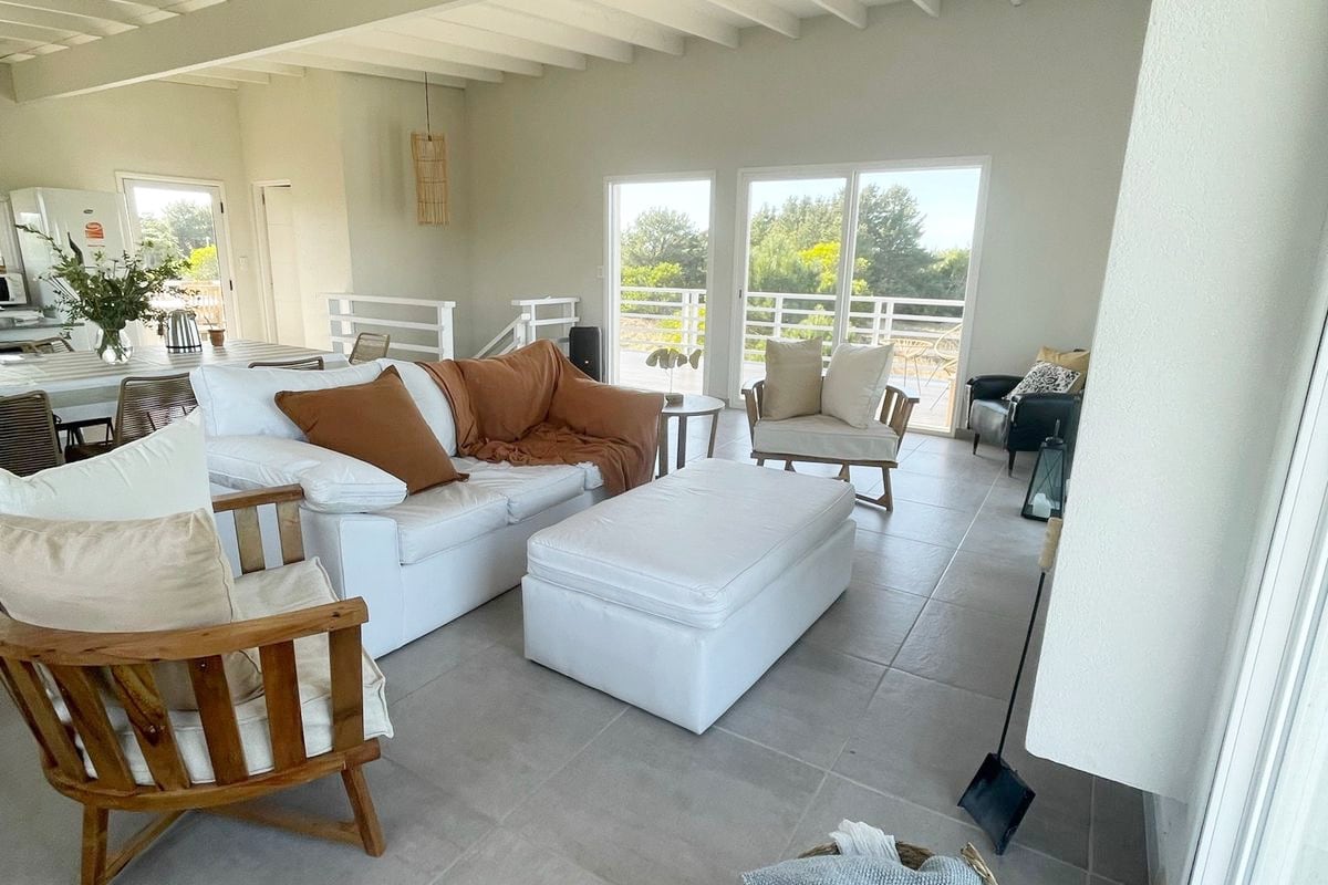 The living area features a combination of comfortable seating, including a spacious white sofa and wooden chairs. Natural light floods the room through expansive windows, showcasing views of greenery. A neutral color palette enhances the inviting atmosphere, complemented by a plush ottoman in the center.