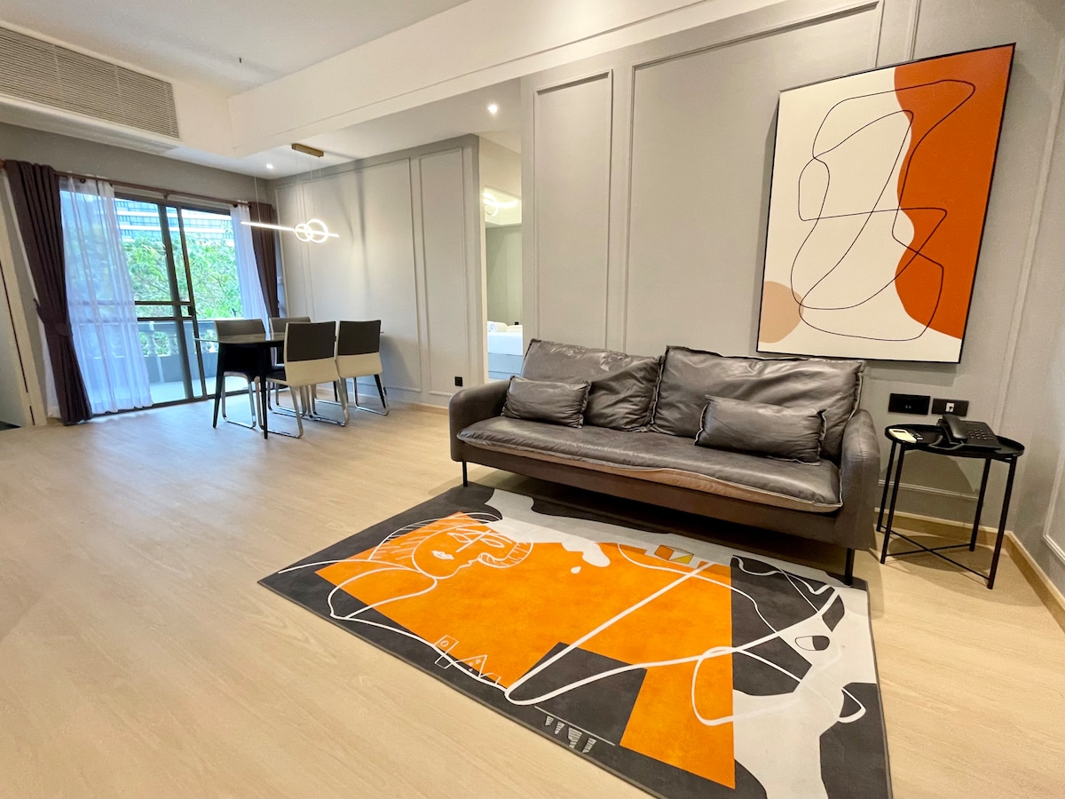 A stylish living area is showcased, featuring a comfortable gray sofa positioned on an abstract orange and white rug. Large glass doors provide natural light and views of the outdoor space. A modern artwork hangs on the wall, complementing the contemporary design.