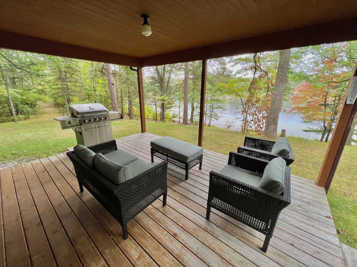 A covered outdoor seating area features four comfortable chairs arranged around a low table, offering views of a tranquil lake and surrounding nature. A grill is positioned nearby on the wooden deck, enhancing opportunities for outdoor dining.