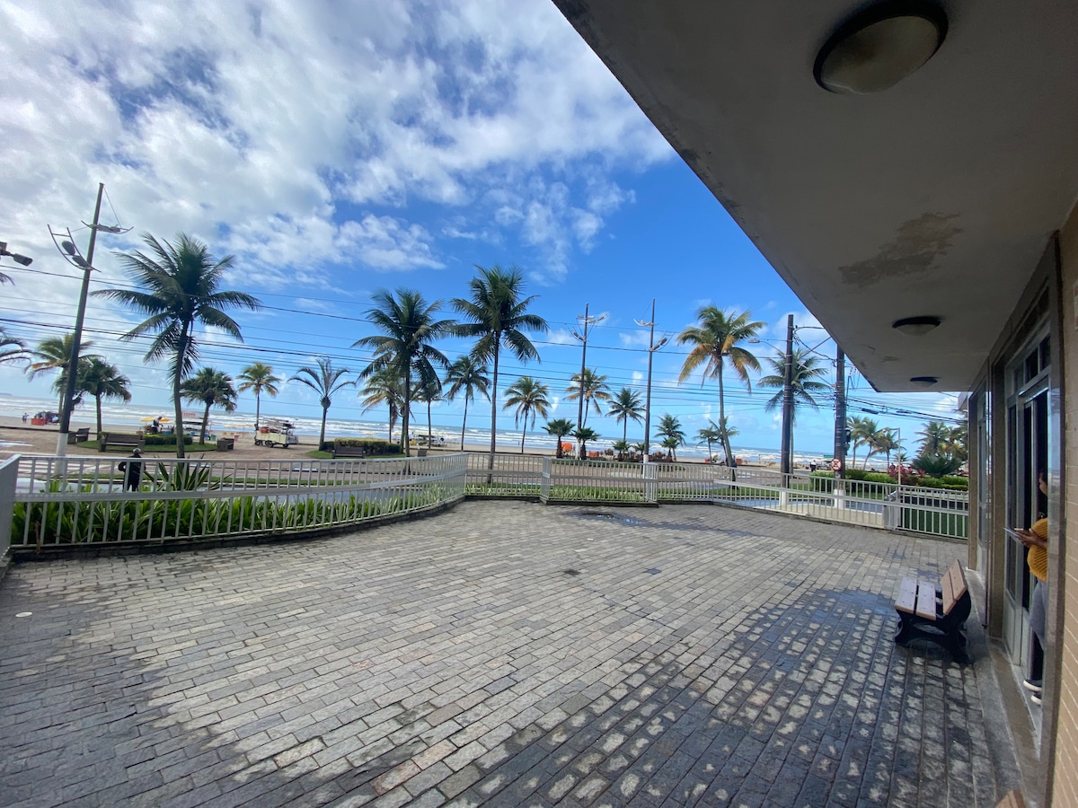 A wide outdoor patio area is presented, featuring a stone floor and several benches for seating. Lush palm trees line the view, with a glimpse of the beach and distant ocean visible. The sky is partly cloudy, allowing for natural light to brighten the space.