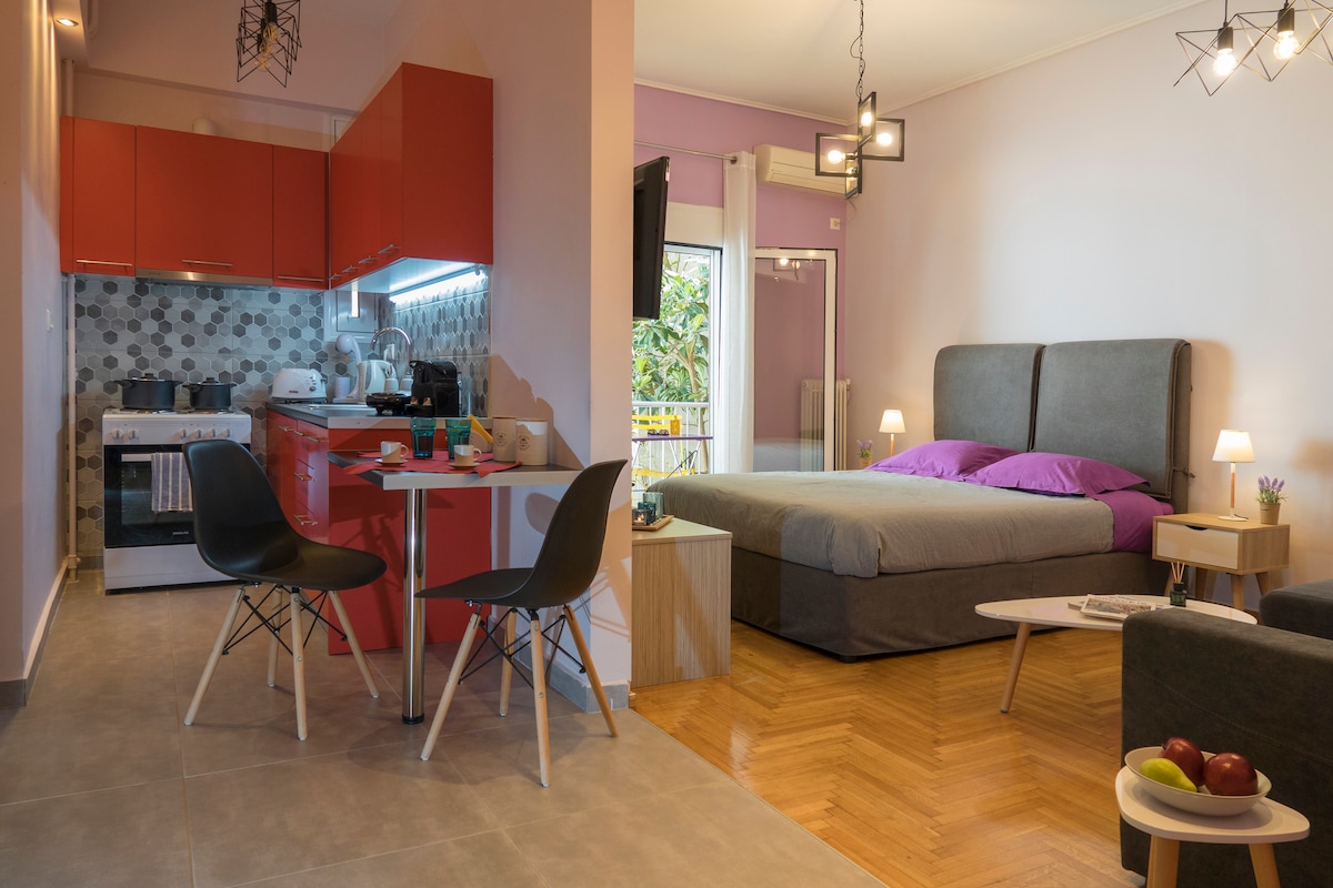 A modern studio layout features an open living area with a spacious bed adorned in purple linens. A small dining table with sleek chairs is situated adjacent to the fully equipped kitchen, which includes bright red cabinets. The room is complemented by natural light streaming in through the balcony doors.