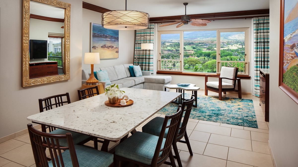 The spacious living and dining area features a marble dining table set for six, surrounded by wooden chairs. A comfortable sofa rests nearby, complemented by a modern armchair. Large windows reveal a scenic view of greenery and hills, while decorative art enhances the room's atmosphere.