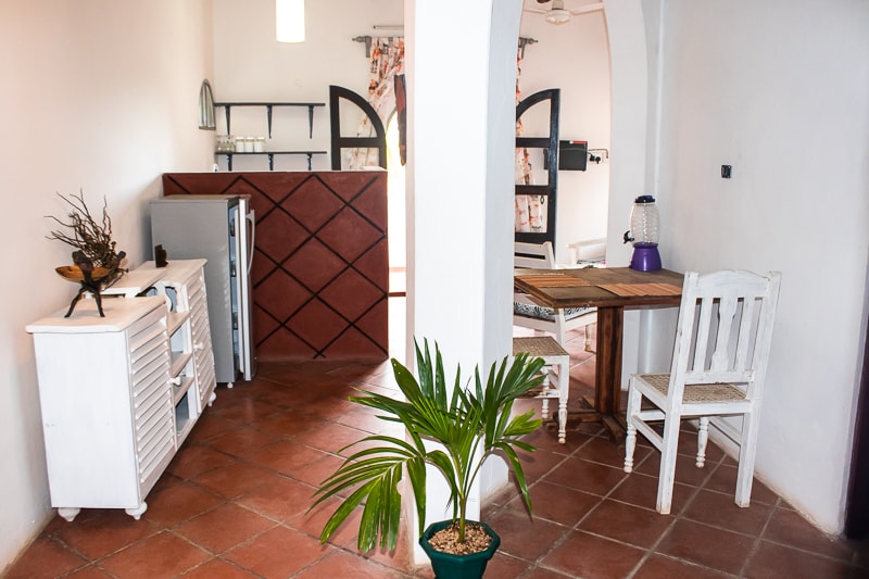 A dining area features a wooden table surrounded by two white chairs, with a small potted plant positioned nearby. A white cabinet and a refrigerator are visible within a cozy, open layout, complemented by warm terracotta tile flooring.