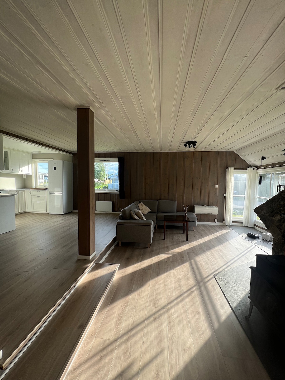 An open living area is depicted, featuring light wooden flooring and a low seating arrangement set against a wall of warm wood panels. Natural light streams through large windows, creating a bright atmosphere. The adjacent kitchen is visible in the background.