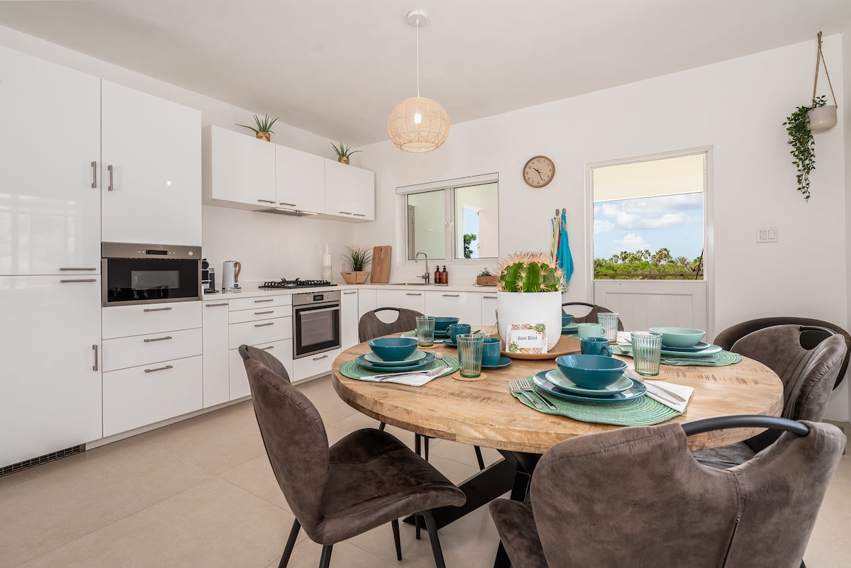 A modern kitchen and dining area feature white cabinetry and a circular wooden table set for six. Natural light fills the space through a window, enhancing the contemporary design. Decorative plants are placed on the walls, adding a touch of greenery to the clean, organized environment.