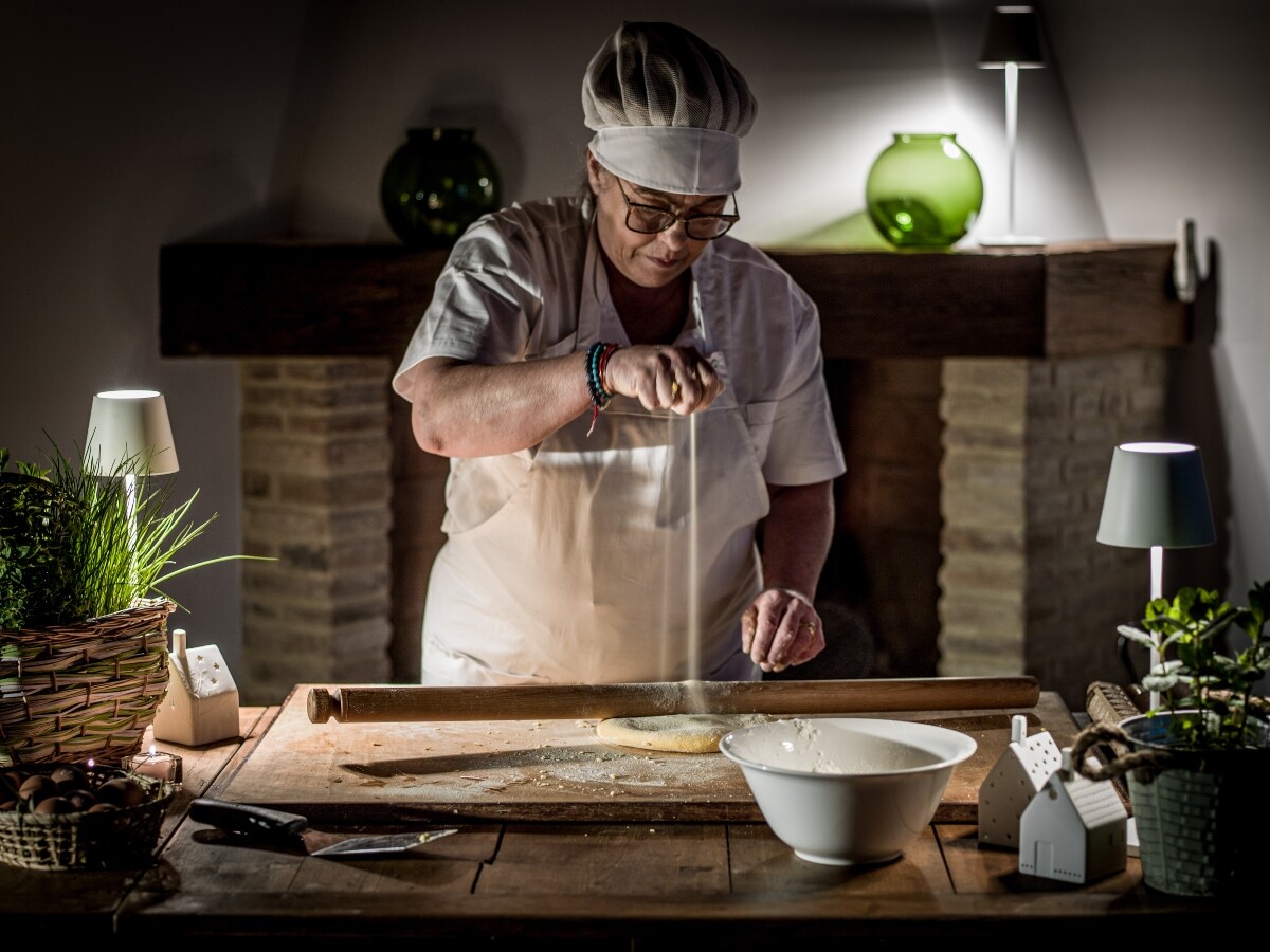 A person is seen preparing food at a wooden table. Flour is being sprinkled over a pastry, with a white bowl nearby containing additional ingredients. Soft lighting is provided by decorative lamps on the table and a green glass accent piece in the background. A stone fireplace adds warmth to the setting.