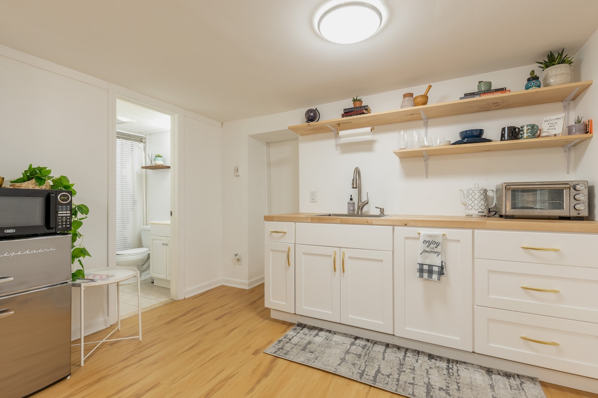 A well-equipped kitchenette features white cabinetry and wooden countertops. Open shelving displays various kitchen items, while a microwave and mini fridge are also present. A soft area rug lies beneath, and a door leads to a bathroom visible in the background.