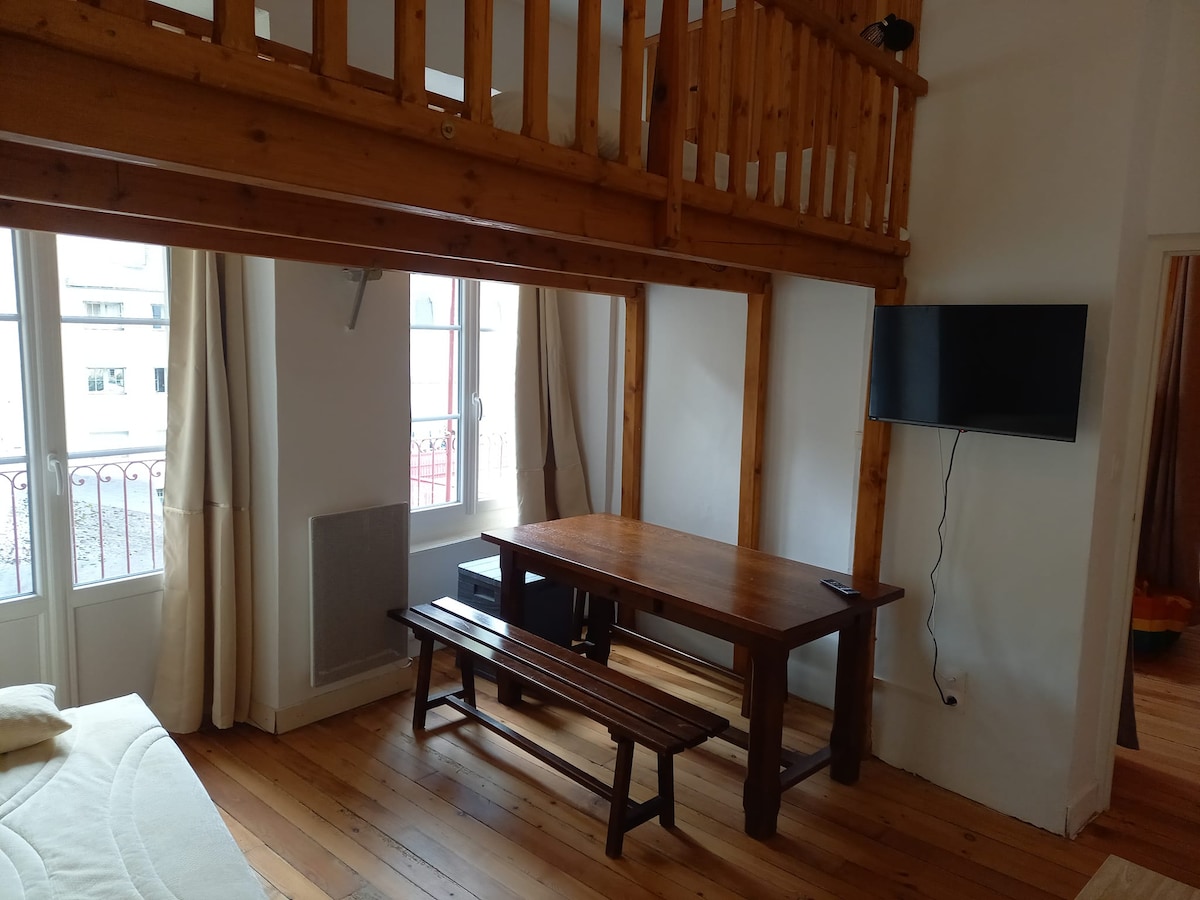 The living area features wooden beams and bright windows, allowing natural light to fill the space. A wooden table is accompanied by a long bench, while a wall-mounted television adds to the functionality. The interior reflects a simple and welcoming design.