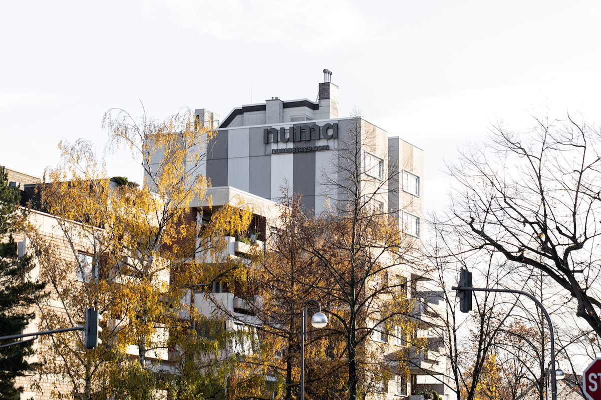 The exterior of the numina building is showcased, featuring a modern facade with a combination of large windows and balconies. Surrounding trees with autumn leaves add vibrant color to the scene, complemented by clear skies above.