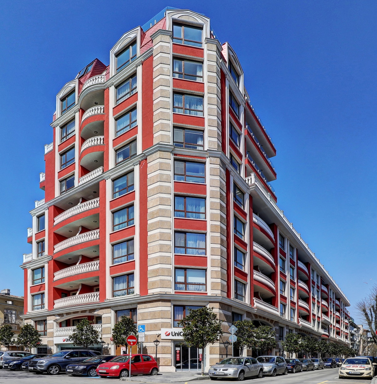 The exterior of a modern building features a striking red facade and multiple balconies. Large windows allow natural light to fill the spaces within. Decorative columns and trees are positioned around the entrance, contributing to the overall architecture in a vibrant urban setting.