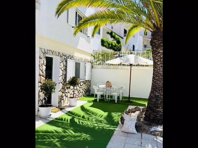 A garden area is presented with green artificial grass and shaded by a large palm tree. A round table with four white chairs is visible under a sun umbrella, alongside potted plants. The backdrop features the white exterior of the house, enhancing the outdoor setting.