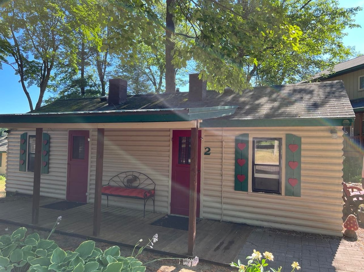 The exterior of Cottage 2 is shown, featuring a log-style facade with green shutters adorned with heart shapes. A small porch, furnished with a bench, invites relaxation. Lush greenery surrounds the cottage, and sunlight filters through the trees, enhancing the welcoming atmosphere.