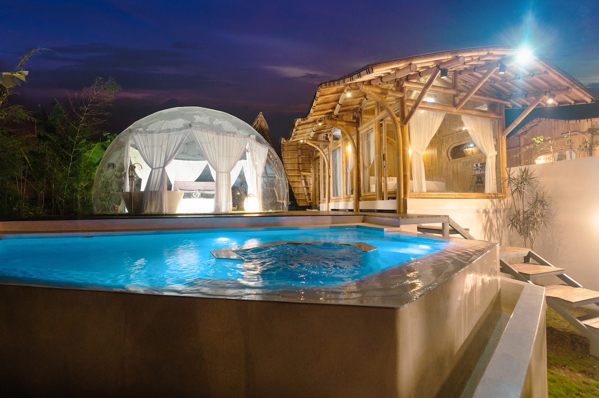 A serene view showcases the glass dome bedroom illuminated at dusk, surrounded by tropical greenery. A spacious private pool reflects the soft lighting, with a bamboo house visible in the background, blending with the natural surroundings. The serene environment emphasizes the peaceful atmosphere.