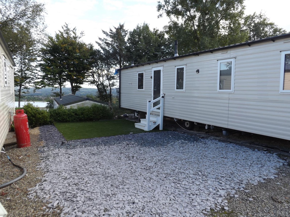 A caravan is positioned on a gravel area, featuring a small patch of green grass at the entrance. Surrounding trees provide natural landscaping, while the view of the reservoir can be seen in the background, contributing to the serene environment.