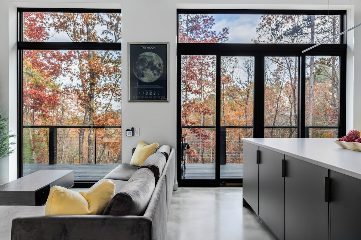 A modern living space features a comfortable gray sofa adorned with yellow accent pillows. Large sliding glass doors provide a view of trees exhibiting vibrant autumn colors. A sleek kitchen island and minimalist cabinetry complete the open-concept design.