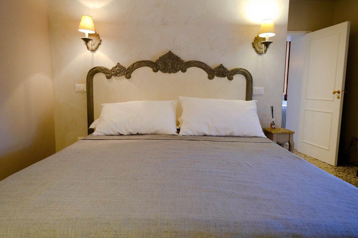 The sleeping area features a spacious bed with a light gray coverlet. Soft white pillows are arranged neatly at the headboard, which is adorned with an ornate design. Gentle lighting is provided by wall sconces on either side.