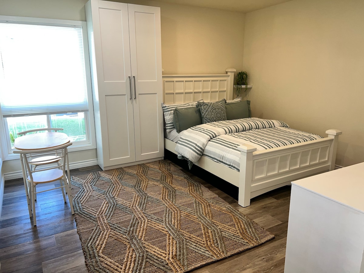 A light-filled studio features a spacious bed adorned with striped bedding and coordinating pillows. A small dining table with two chairs sits nearby, complemented by a textured area rug. A tall white wardrobe provides storage, while natural light filters through the window.