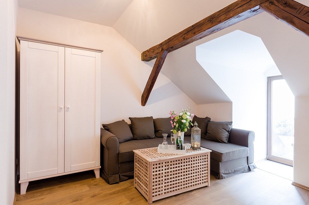 A cozy sitting area features a light-colored cabinet and a comfortable sofa with neutral cushions. A coffee table with a woven design holds decorative items, while natural light pours in through a large window, illuminating the space warmly.