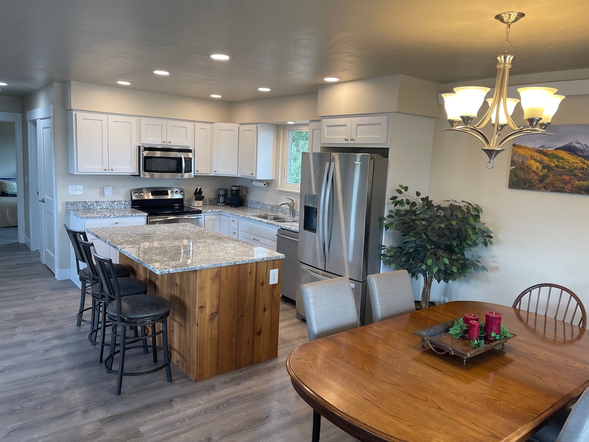 An open-concept kitchen and dining area feature modern appliances and a large island with seating. A wooden dining table accommodates several chairs, while natural light highlights the neutral color palette. Notable elements include a chandelier above the table and a scenic wall-mounted artwork.