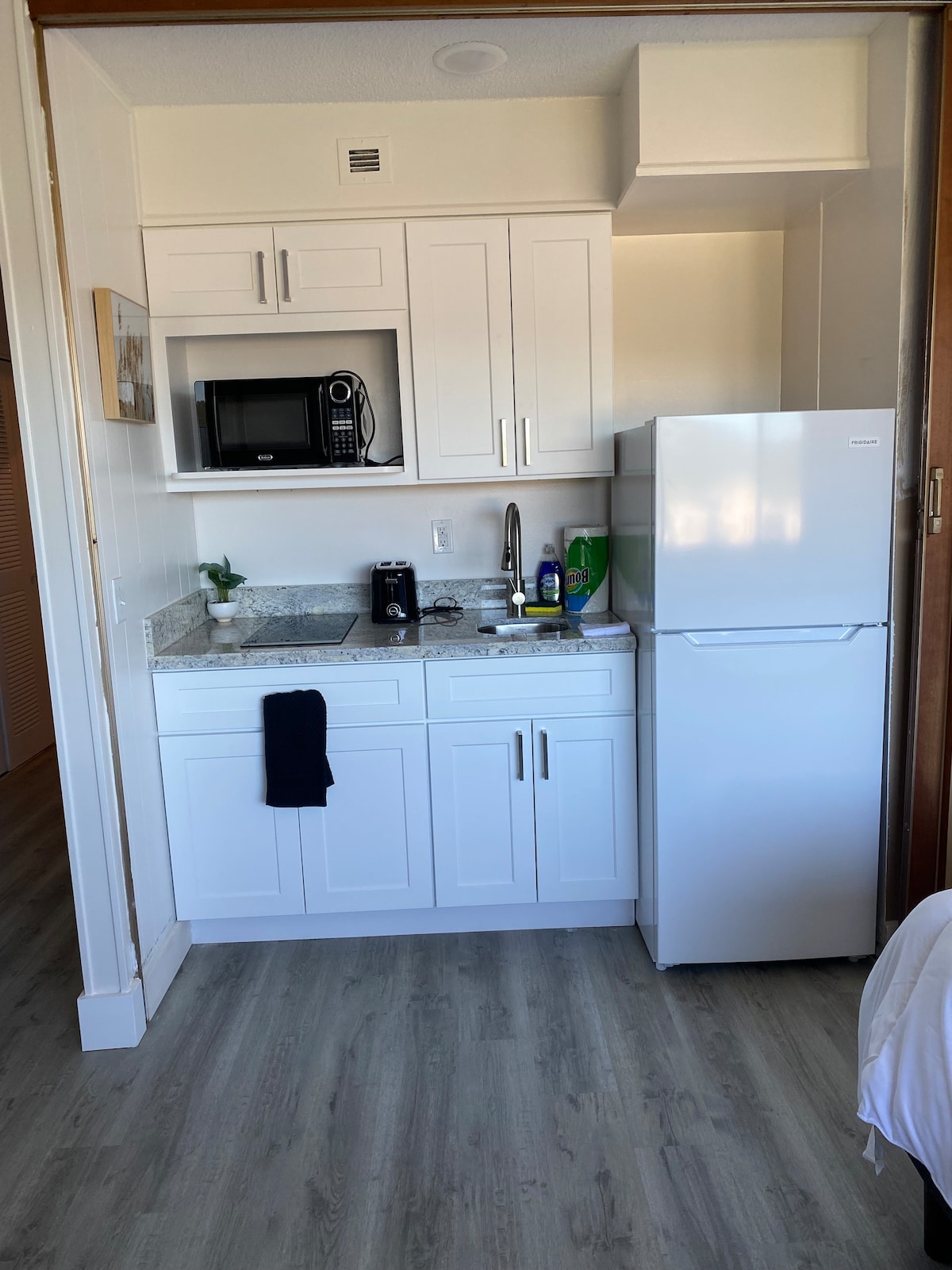 A compact kitchen area features modern cabinetry and a granite countertop. A microwave and refrigerator are visible, along with a sink and some kitchen essentials. Natural light illuminates the space, enhancing its functional layout.