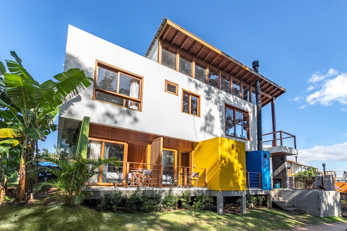 The two-story house features a modern design with large windows allowing natural light to illuminate the interior. A vibrant yellow accent wall contrasts with the white exterior, while spacious balconies overlook the surrounding greenery. The inviting outdoor area includes manicured grass and tropical plants.