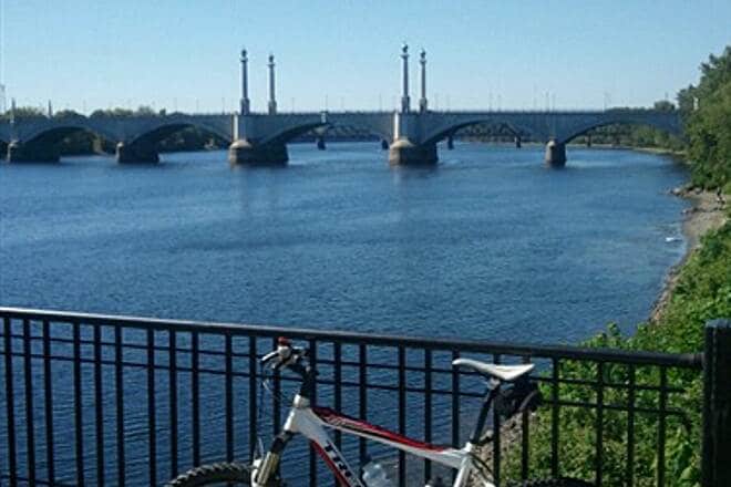 A view of the Connecticut River showcases a tranquil water scene, framed by vibrant greenery along the bank. A bridge with tall pillars is visible in the background, while a bicycle is parked against a decorative black railing in the foreground.