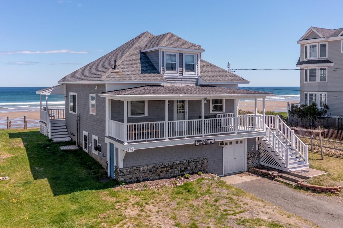 The exterior of a New England cottage is shown, featuring a gray facade and a spacious wrap-around porch. The sandy beach and ocean are visible in the background, enhancing the coastal setting. The grassy area surrounds the house, leading to a private driveway.