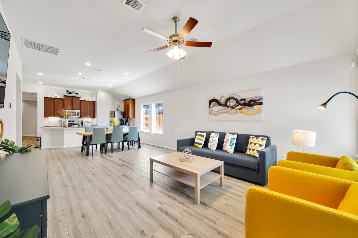 A bright open living area features a gray sofa adorned with patterned pillows, complemented by two yellow armchairs. A light wood coffee table sits in the center, with a ceiling fan above. The kitchen is visible in the background, equipped with wooden cabinetry and a dining space.