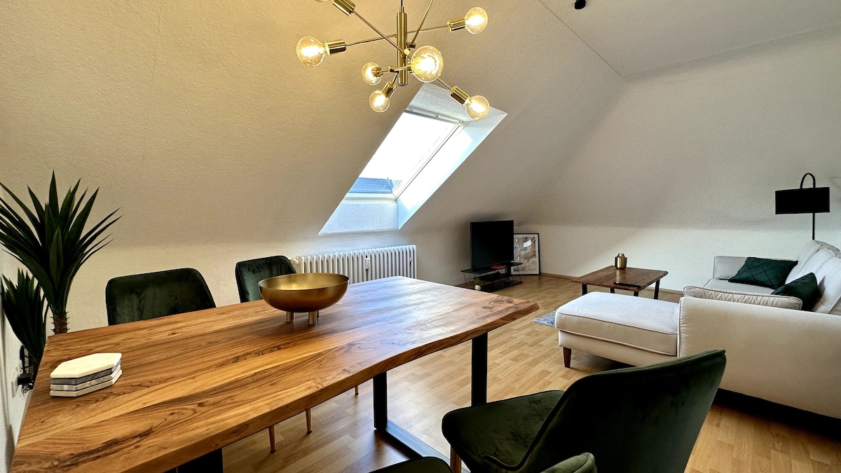 A spacious living area is highlighted by a wooden dining table accompanied by dark green chairs. A stylish chandelier hangs from the ceiling. A comfortable sofa is positioned next to a flat-screen TV, while a large skylight illuminates the room.