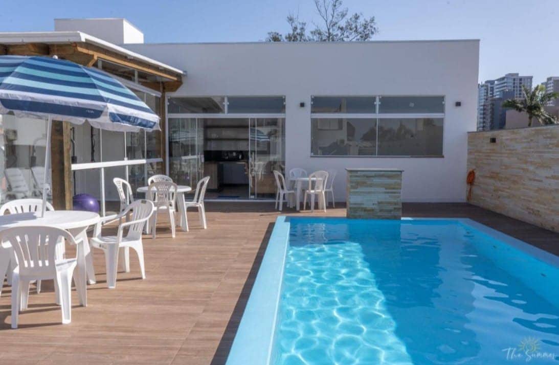 A swimming pool is situated beside a wooden deck, with two tables and multiple chairs arranged nearby. An umbrella provides shade over one of the tables. Large glass doors offer a view into the kitchen area, contributing to a bright and open atmosphere.