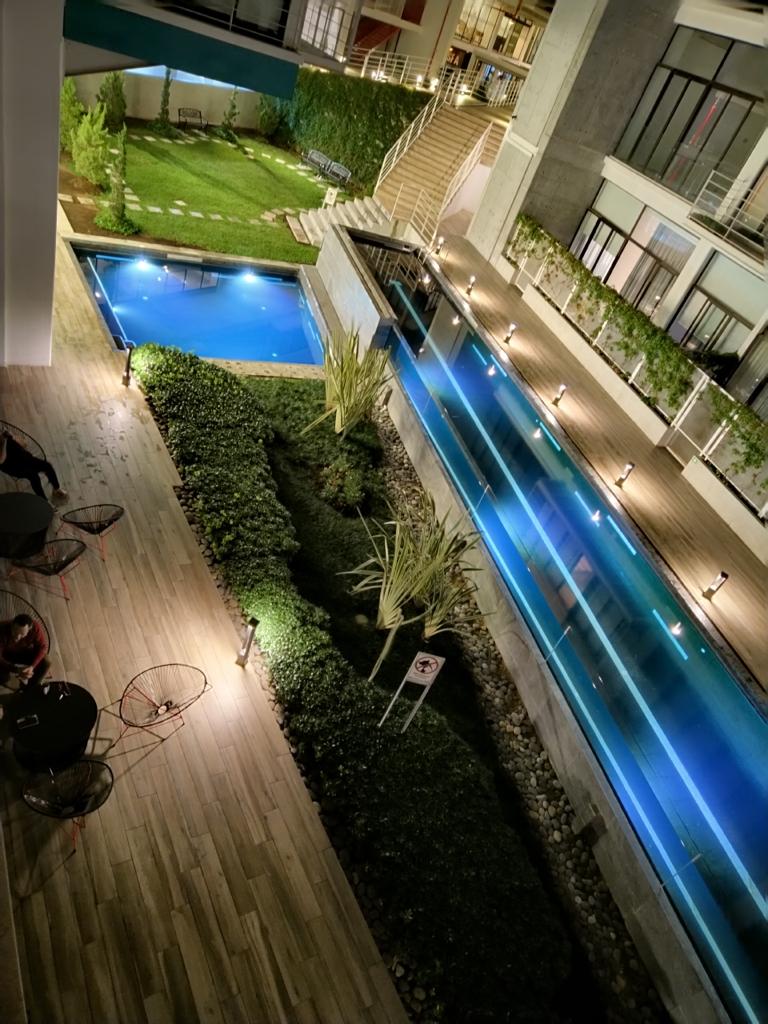 An outdoor area is presented, featuring a lap pool with illuminated blue water. Surrounding greenery and a well-maintained lawn are visible, along with pathways lined by soft lighting. Seating areas with chairs are arranged nearby, contributing to a serene environment.