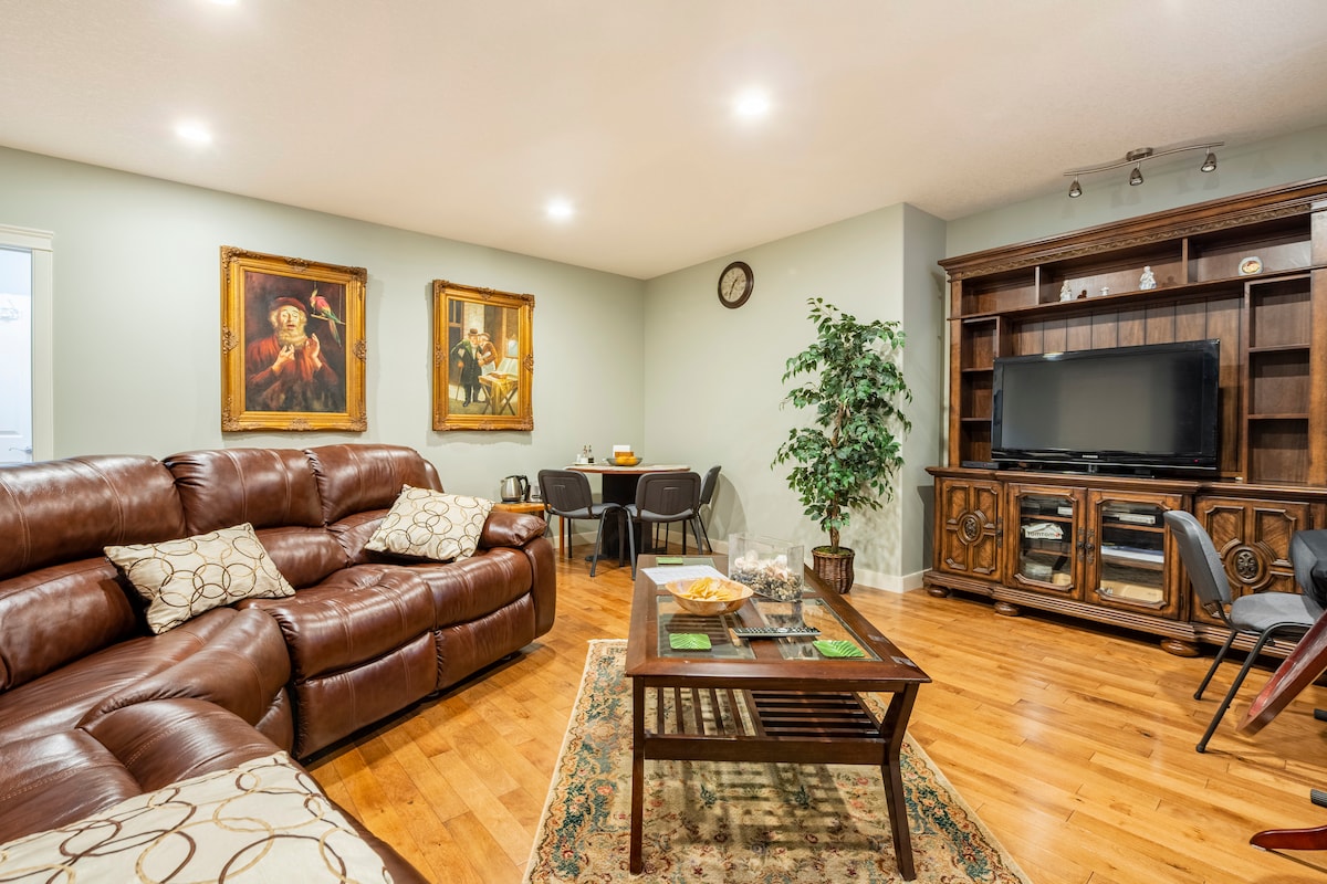 A spacious living room is furnished with a large brown sectional sofa, complemented by a wooden coffee table on a decorative rug. Natural light enhances the ambiance, while two framed paintings adorn the walls. A television is integrated into an elegant wooden unit, with a dining area visible in the background.
