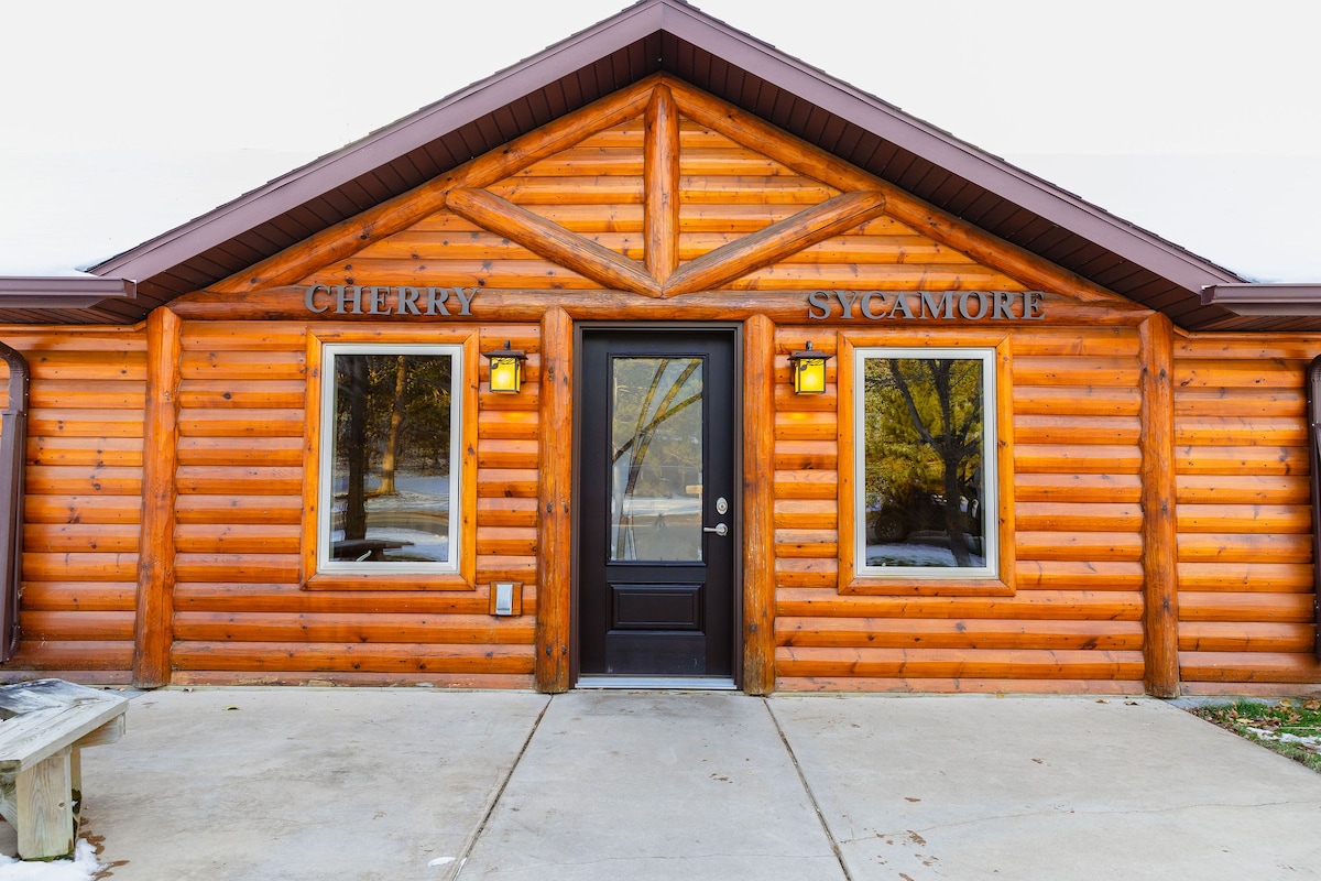 The exterior of the cabin features a log-style design with warm wooden tones. Two large windows frame the entrance, which is adorned with a black door and rustic lanterns. The name 'Cherry Sycamore' is displayed prominently above the windows.