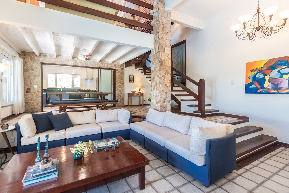 A spacious living room features a large sectional sofa in neutral tones with blue accents. A wooden coffee table sits in front, while a striking piece of wall art adds color to the scene. Natural light floods the area through windows, highlighting the stone accents and staircase.
