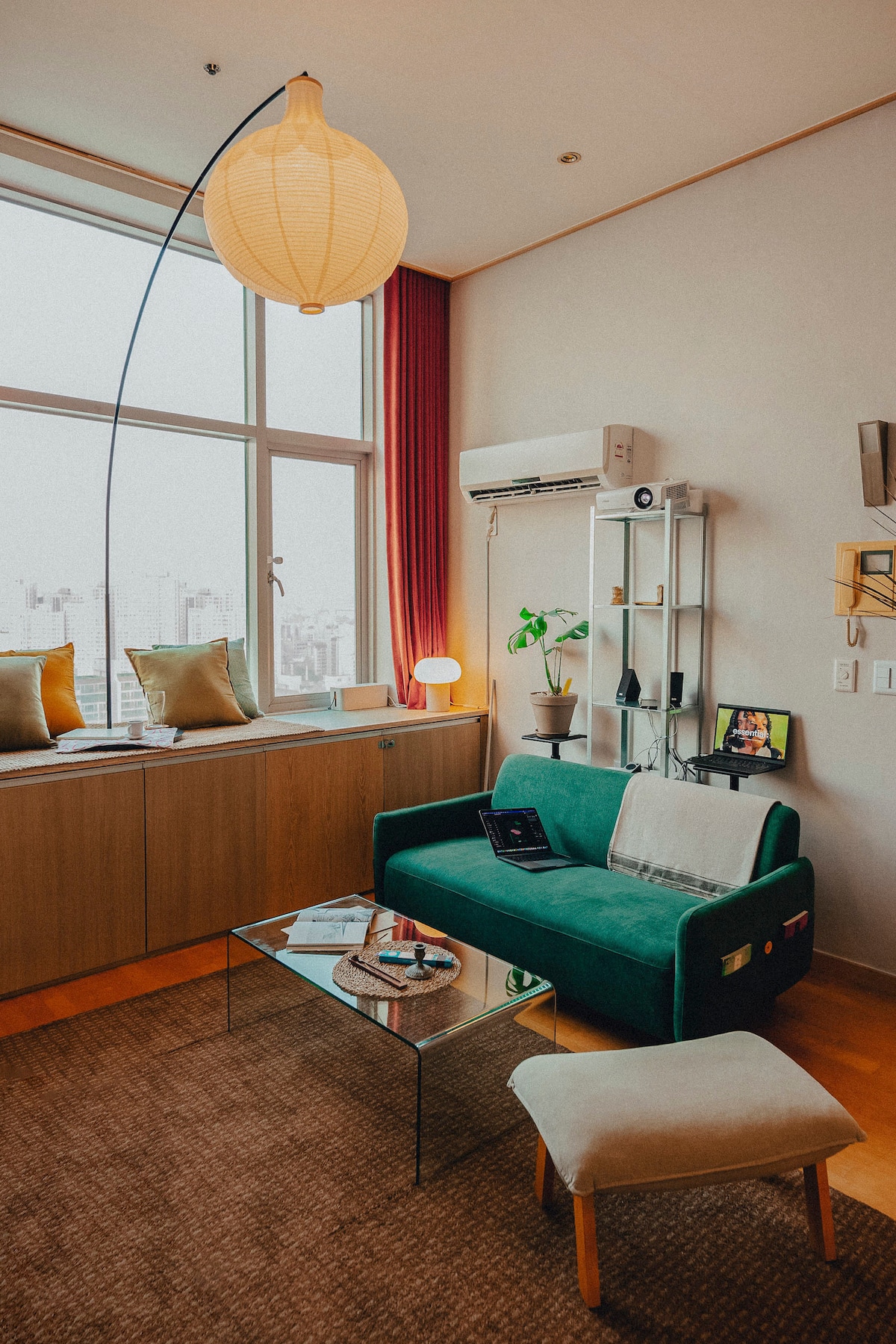 A comfortable seating area showcases a green sofa paired with a light-colored ottoman. A glass coffee table sits in front, complemented by decorative items. Large windows provide ample natural light, while a floor lamp and potted plant add a touch of warmth to the space.
