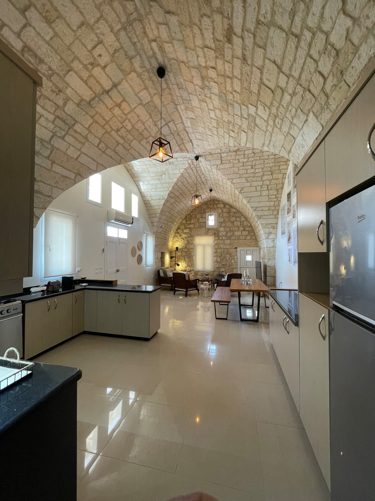 The kitchen area features high arched stone ceilings and sleek cabinets, creating a blend of traditional and modern design. Large windows allow natural light to illuminate the spacious room. A dining area is visible in the background, providing a welcoming space for gatherings.