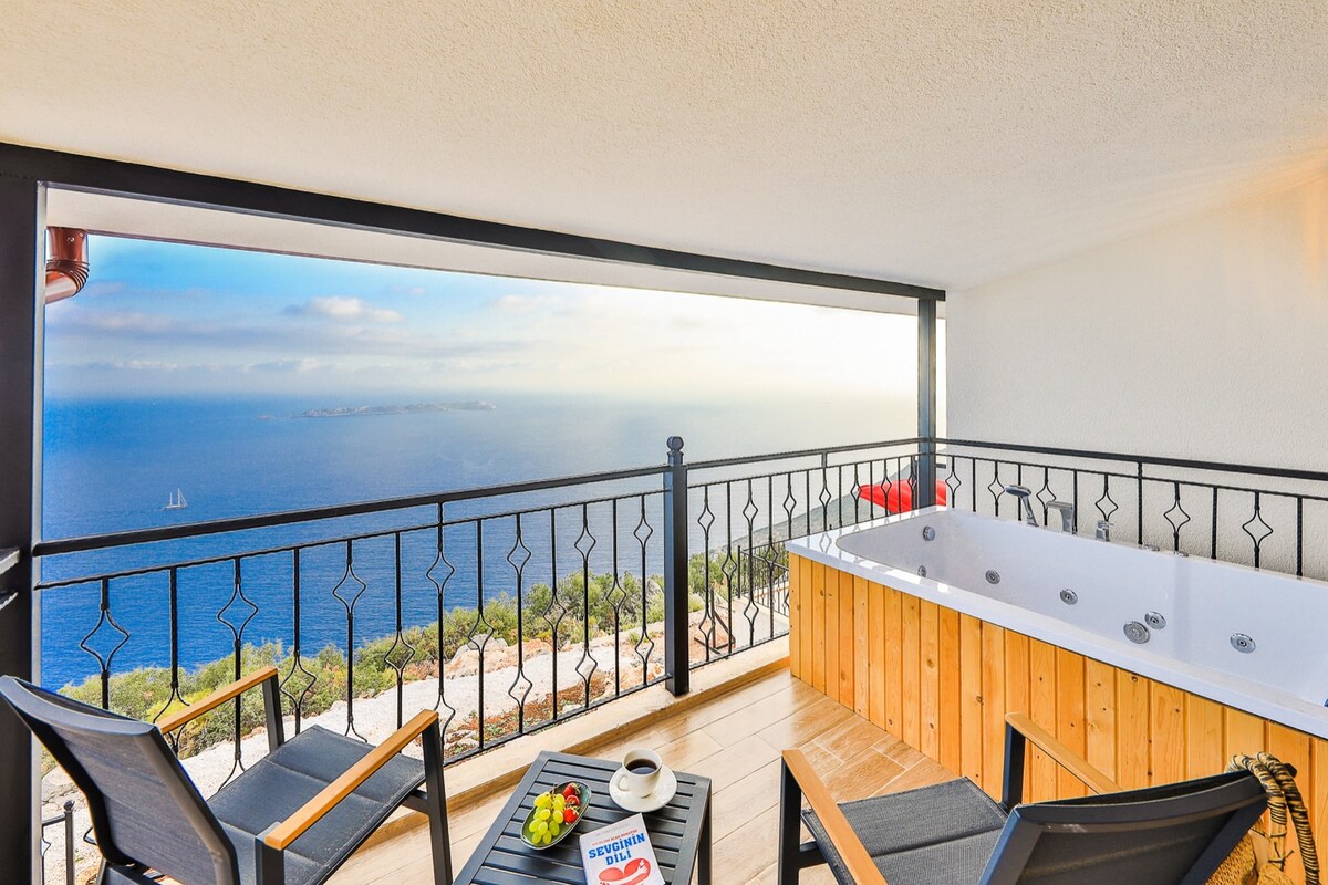 An outdoor terrace offers a view of the serene ocean, featuring a wooden hot tub with jets. Two black chairs are positioned around a small table, which holds a bowl of fruit and a magazine. The space is framed by a decorative black railing.