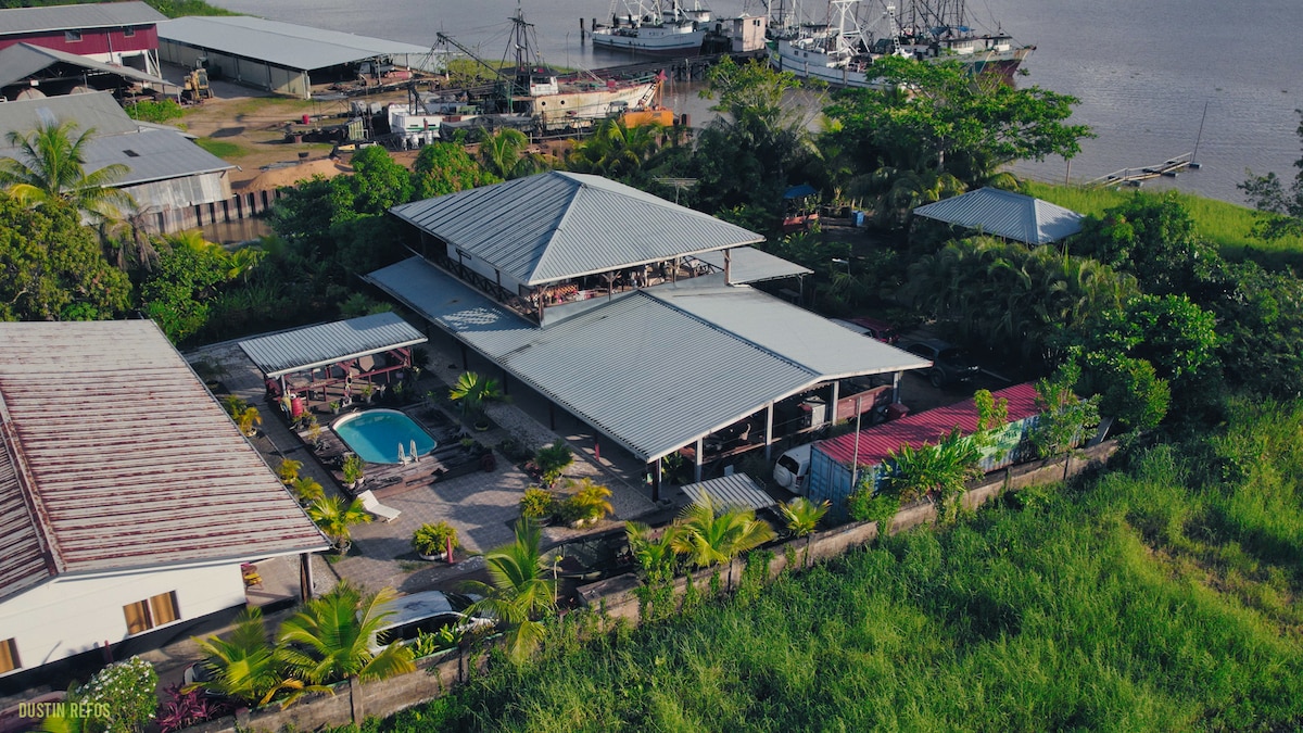 An aerial view captures the charming property surrounded by lush greenery. The pool is centrally located, with patio seating visible. Nearby structures include additional apartments and a view of the river and boats, enhancing the tranquil ambiance of the space.