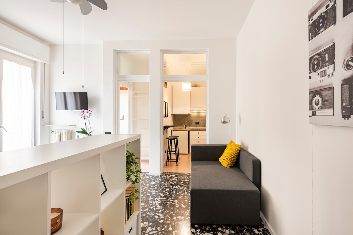 A modern living space is presented with a soft gray sofa and a bright yellow pillow. A wall-mounted television is positioned opposite. Natural light filters through the windows, highlighting the glossy, patterned floor. A sleek bookshelf and a small dining area are subtly included in the open layout.