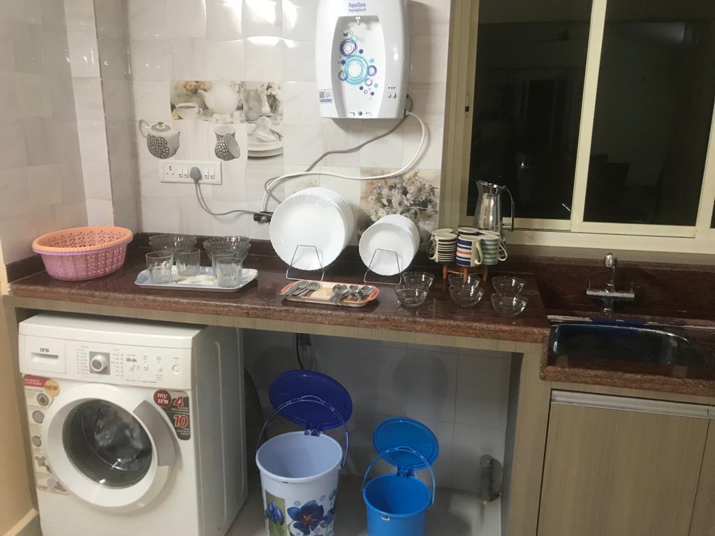A functional kitchen area features a washing machine, various dishes, and glassware neatly arranged on a countertop. Colorful buckets are positioned below, with a sink alongside. Natural light enters through a nearby window, enhancing the workspace's utility.