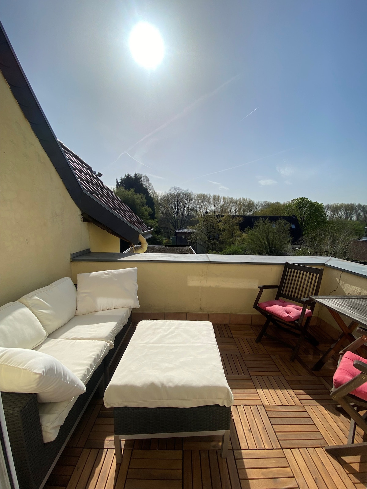An outdoor terrace is showcased, featuring a comfortable seating area with plush white cushions and a matching coffee table. The space is surrounded by a railing and offers views of green trees and a clear sky. Sunlight illuminates the area, highlighting the wooden deck.