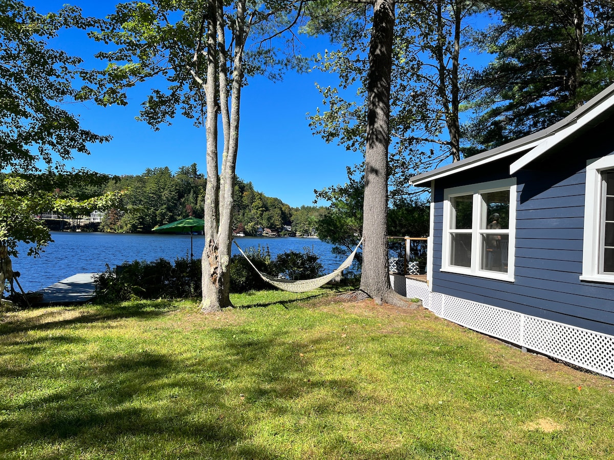 A spacious grassy yard is bordered by mature trees, with a hammock gently swaying between two trunks. The serene waters of Horace Lake are visible in the background, framed by lush greenery and a clear blue sky above.