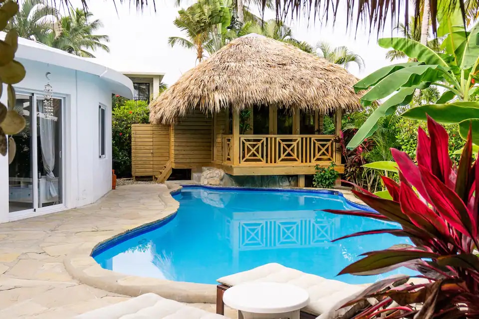 A clear blue swimming pool is bordered by natural stone, leading to a thatched-roof gazebo. The gazebo features wooden railings and a dining table. Lush tropical plants surround the area, adding vibrant greenery to the scene.