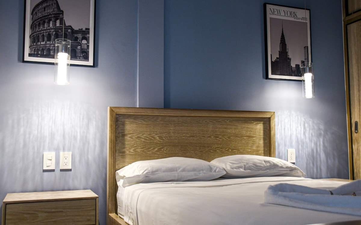 A modern sleeping area is presented with a spacious bed and two nightstands. Framed artwork depicting cityscapes adorns the blue walls. Soft lighting from mounted sconces enhances the serene ambiance, contributing to a calming environment.