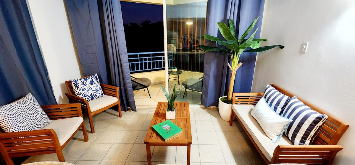 A welcoming living area features wooden seating arrangements with cushions, complemented by a coffee table with a green book. A large window is adorned with dark curtains, providing a view of the outdoors. A potted plant adds a touch of greenery to the space.