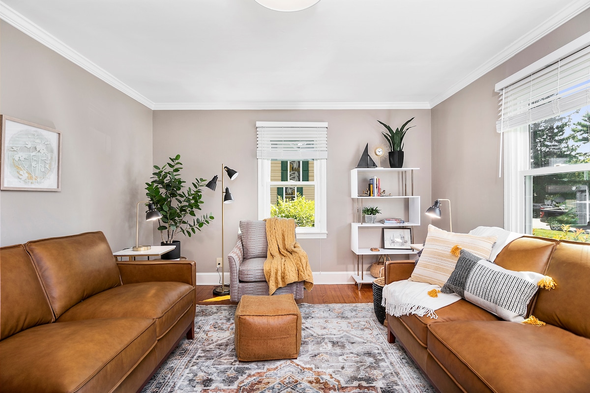 A cozy living area features two brown leather sofas, a patterned rug, and a soft ottoman. A reading nook is created with a gray chair draped in a yellow blanket. Natural light streams in through a large window, with plants and bookshelves enhancing the space.