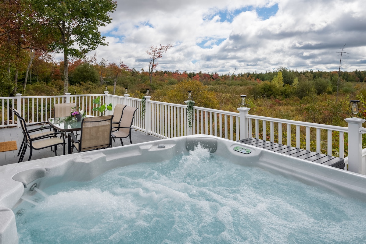 A relaxing hot tub is positioned on a spacious deck overlooking vibrant foliage and natural greenery. Nearby, a dining table with chairs can be seen, enhancing the outdoor space for gatherings. The sky is partially cloudy, adding to the serene atmosphere of the retreat.